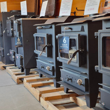 Stoves & FireplacesA row of black wood stoves is displayed on wooden pallets in a showroom. Each stove has visible knobs and handles, conveying a practical and industrial tone.