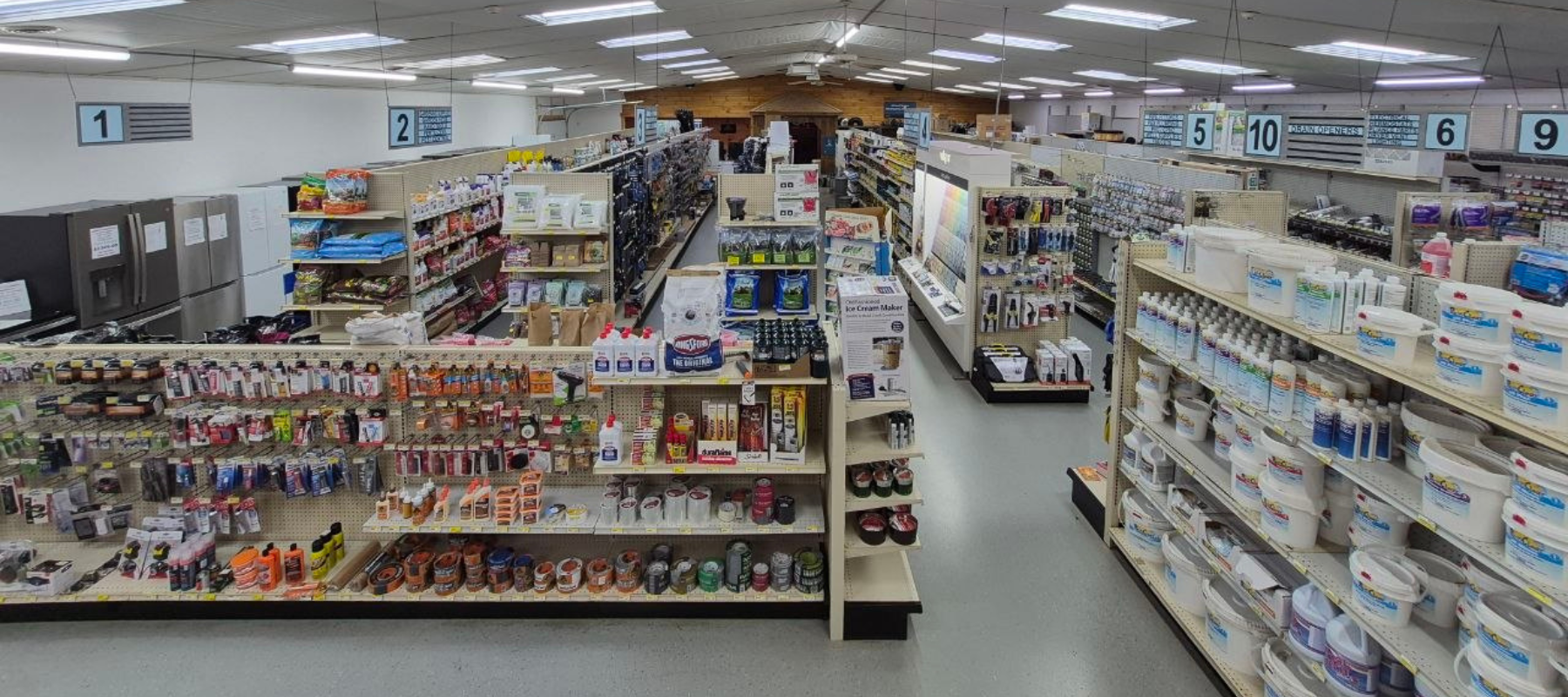 Wide view of a hardware store interior featuring neatly arranged aisles filled with tools, paint, cleaning supplies, and household items. Bright lighting.